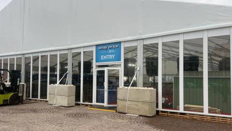 Jersey on Ice ice rink at St Helier entrance. A big white marquee is covering the ice rink. Transparent windows are all around the marquee. A large blue entry sign is above the door.