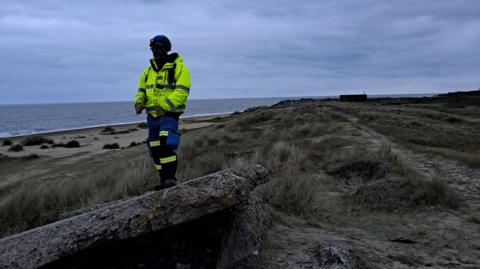 A person in a high vis yellow jacket is standing on a piece of rock next to the coast. The sea and beach can be seen in the background and it appears to be the early morning. The persons face is not visible. 