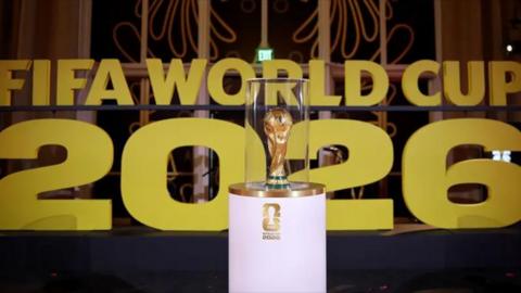 The FIFA World Cup trophy inside a case on a podium infront of a sign naming the 2026 tournament.