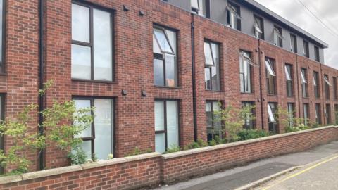 A view of Valley Court in Sandbach, a modern red-brick building with a second storey in black cladding, set back from the ground and first floors. Trees and bushes are sprouting between the building and the boundary wall onto the street. 