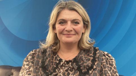 Headshot of Bryony Gordon - a woman with shoulder length blonde hair and wearing a leopard print top. She is smiling and there is a blue background behind her.