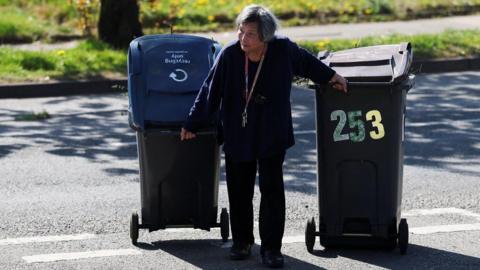 A woman pulls her wheeled bins to a Mobile Household Waste Centre where residents can dispose of their rubbish. She is wearing a dark top and trousers and has a lanyard around her neck. She is pulling two bins and stooping slightly. The sun is out and there is grass behind her and a tree trunk.
