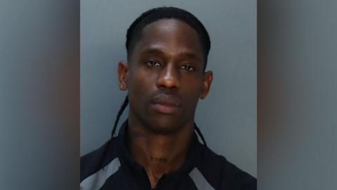 A mugshot of Travis Scott, his hair tied back. His expression is blank as he looks into the camera. He appears to be wearing a black and silver tracksuit top.