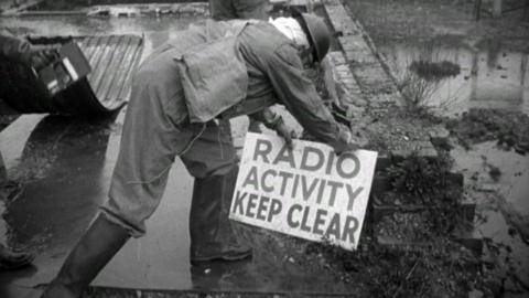 Black and white image of a man in protective clothing of the time, placing a sign that reads 'RADIO ACTIVITY KEEP CLEAR' in a wasteland during a Nuclear Bomb drill.