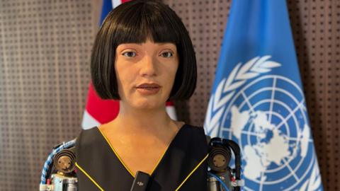 The ultra-realistic humanoid robot artist Ai-Da speaking to the camera. The United Nations flag is behind her.