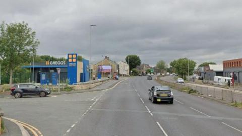 A photo of a road in Bradford which is a dual carriageway with a Greggs on one side and a few black cars on the road 