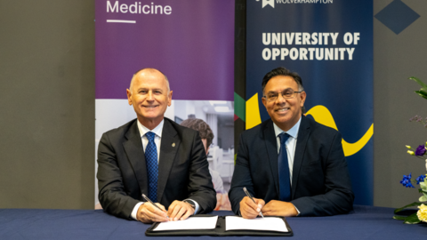 Two men sit at a table with a blue tablecloth and paperwork in front of them with pens in their hands. They have two banners behind them. A purple one behind Subic is from Aston with a picture on it. A blue banner is behind Adia with the University of Wolverhampton insignia and the words University of Opportunity. An arrangement of flowers is visible to the right of the men.