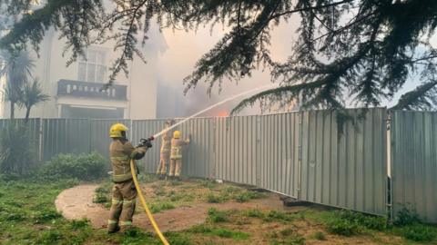 A firefighter aims water from a hose over a high metal fence. On the other side of the fence can be seen buildings, flames and smoke.