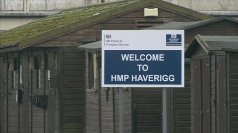 A blue and white sign saying "Welcome to HMP Haverigg". It is in front of hut-style buildings painted brown outside the main gate of the prison.