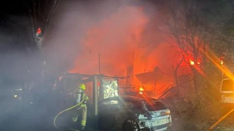 The orange glow of a fire, and smoke billowing from it, can be seen in the background at night time. In the foreground is a firefighter, with his back to the camera and wearing the brown and yellow uniform. There is a damage car in the foreground too.