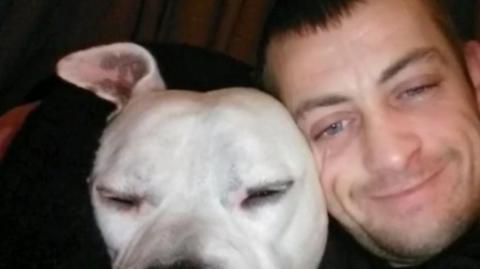 A man with short brown hair and stubble is next to a white Staffordshire bull terrier-type dog.