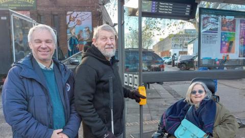 Two men stand by a bus stop alongside a woman using a wheelchair. All three are smiling towards the camera. The man in the middle is holding a walking stick and has his other hand on a large yellow button attached to the side of the bus shelter.