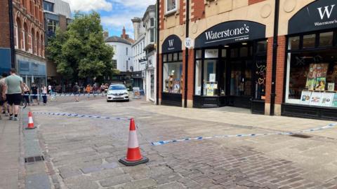 Blue police tap attached to cones and a building has cordoned off a section of a city street in Norwich, outside a Waterstone's store, which has black and white branding, with books in the windows on display. A police car can be seen in the background next to the cordon, as people walk past it. 