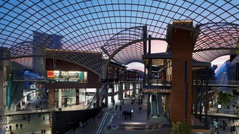 Multi-tier shopping centre with large glass roof arcing between the different buildings. Three avenues on different levels are visible. People can be seen walking down the avenues, and an escalator can be seen in the middle. The sky is blue and pink at sunset.