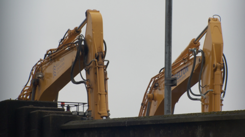 Two yellow diggers pictured behind a wall against a grey sky