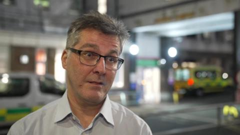 Dr James Marsh, a middle aged man with grey hair glasses standing outside a hospital