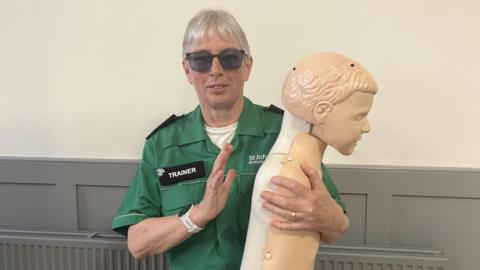 A woman wearing glasses and a green St John Ambulance shirt is holding a mannequin facing to the side. She has a badge on her shirt that reads TRAINER.