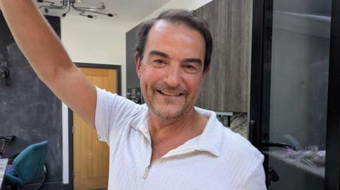 Adrian Collis stood in his house smiling for the camera. He has dark hair with stubble and has his arm up resting his had on a wall. He has a white polo-style half-zip shirt on.