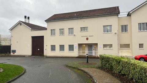 A yellow building with bars on the windows and a logo saying Guernsey Prison above it. 