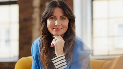 A woman with long brown hair and a blue suit, she is sitting on a yellow sofa and is resting wit her hand on her chin