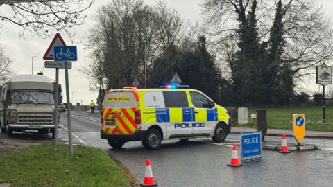 A police van parked across a road which has grass and pavement each side. Cones are also placed across the road and a blue sign to say the road is closed.