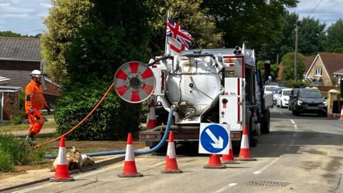 A white tanker truck is parked next to a green hedge. There is a row of cars on the opposite side of the road. There are orange cones behind the vehicle and a man wearing orange florescent overalls. There are pipes running from the read of the tanker.