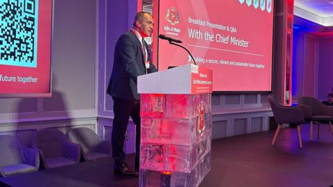 Chief Minister Alfred Cannan stands at a lectern delivering a speech. He is wearing a dark suit and is surrounded by large screens with red displays with the government crest and the words "Breakfast Presentation & Q&A with the Chief Minister".