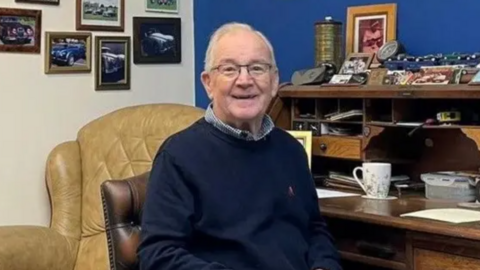 A man with grey hair and glasses is sat at a desk, smiling to the camera. Behind him are pictures of old cars