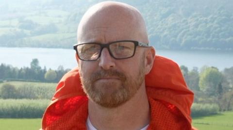 Matt Cox looks at the camera. He is bald with a ginger beard and glasses. Behind him is a large lake and trees