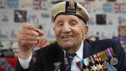 Dougie Shelley wearing his suit jacket with a row of medals pinned to his chest. He is wearing a white shirt and navy tie. He also wears a white naval beret and is holding a small glass up in toast.