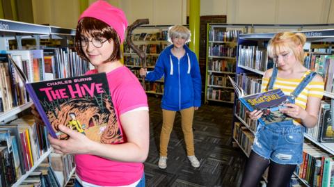 Three young people stand in a library wearing outfits inspired by characters from graphic novels. One holds a title called The Hive, by Charles Burns. Another holds a graphic novel called Sugar Skull, by the same author. The third person in the background is holding a large wooden cane. 