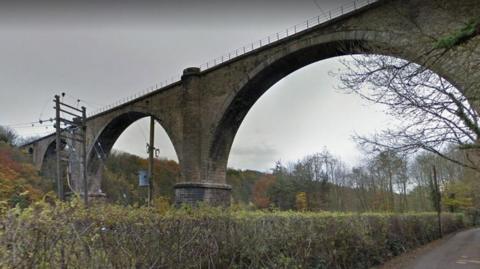 The stone Victoria Viaduct photographed from the path below. The viaduct has fencing with trees and shrubs surrounding its foundations.