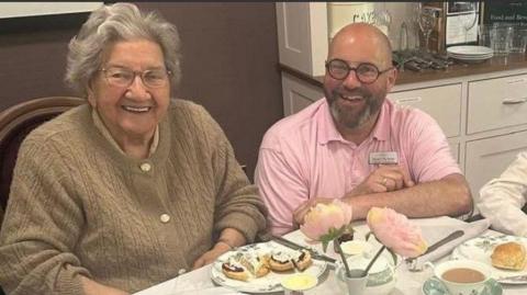 A square table covered in a white table cloth, with a tray of jam, cream and scones, tea cups containing tea and plates with scones on. An elderly woman is sitting to the left with medium-length grey hair and a brown cardigan, Martin has very short hair and a brown beard and glasses and is wearing a pink shirt, 