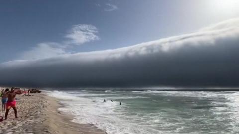 People on a beach are looking at a huge roll cloud over the ocean