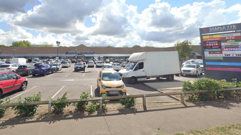 A shopping centre car park. There are several cars parked and a white van is driving through the car park. On the right is a sign with the words "Staple Tye Shopping Centre" above the logos of 18 businesses.