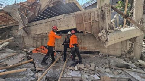 Rescuers clad in bright orange long sleeved vests and helmets inspect the rubble of a collapsed building at an Islamic boarding school in Sidoarjo, East Java, Indonesia. They are standing in front of a huge heap of twisted concrete and rebar.
