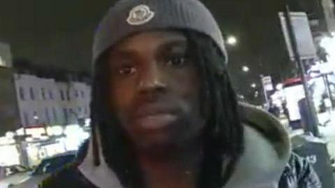 A close-up, slightly low-angle photo of a Jordan Asije, seen with dreadlocks outdoors at night. He is wearing a grey beanie, a grey hoodie, and a dark jacket while looking directly into the camera on a city street.