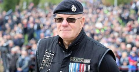 Martin Dickinson, wearing a black beret and a black gilet with medals hanging on his left breast. He is standing in front of a large crowd of people.