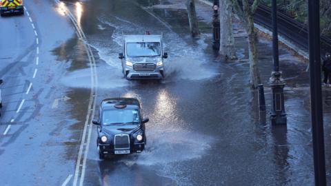 A black cab and a van driving through floodwater in Victoria Embankment in central London