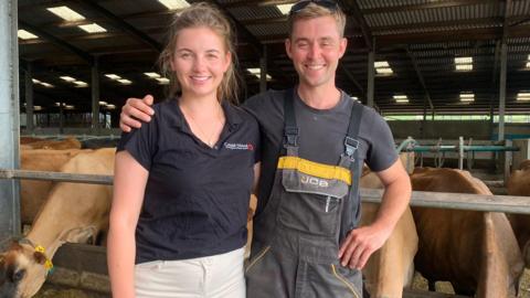 The picture shows two individuals standing inside a barn or livestock facility. 
One person is wearing a black polo shirt and white pants, holding sunglasses.
The other is dressed in grey overalls with a black t-shirt underneath.
The barn has a metal roof with skylights that let in natural light.
Several cows are visible in the background, eating from feeding troughs.