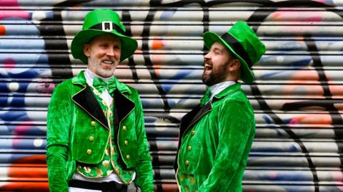 Two men, dressed like leprechauns, are standing having a laugh. The backdrop is a shop shutter that is covered in graffiti