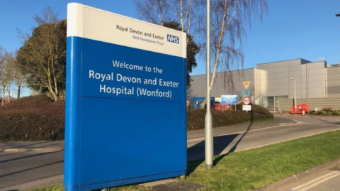 A blue sign which says 'Welcome to the Royal Devon and Exeter Hospital (Wonford)'. The hospital sits behind it