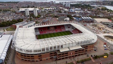 An aerial shot of the Stadium of Light which is a large, rectangular, open stadium with a white roof above the stands. There is no roof above the pitch and Sunderland badge can be seen picked out in the seating. The club name is spelled out in white seating among the otherwise red seats. Around the stadium is a lot of empty land with houses and flats in the distance.
