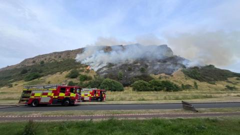 Fire trucks parked at foot of hill which is on fire