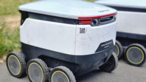 A small delivery robot. It is white and grey and less than a meter high and has six wheels. It is parked next to another robot.