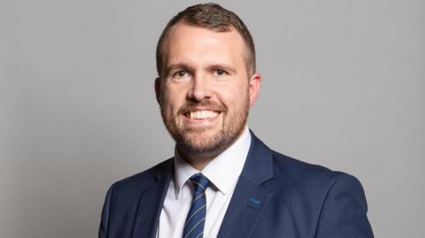 Jonathan Gullis - a man with brown hair, a beard, a blue suit jacket, blue striped tie and white shirt - smiles while in front of a grey wall.