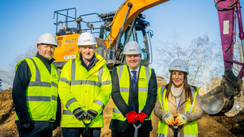 Three men and woman are stood in front of a digger. The ground-breaking has begun. All four people are wearing white hard-hats and yellow hi-vis jackets.