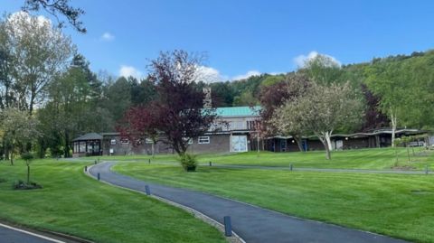 A crematorium building located on large grassed area with trees in the foreground and background. There is a small road running up to the building. 