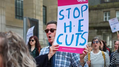 A man in sunglasses and a checked shirt holds a sign which says: Stop the Cuts