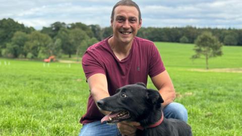 A man in a red T-shirt and denim jeans, smiling widely, he is squated down over a black dog, on green grass with forestry in background